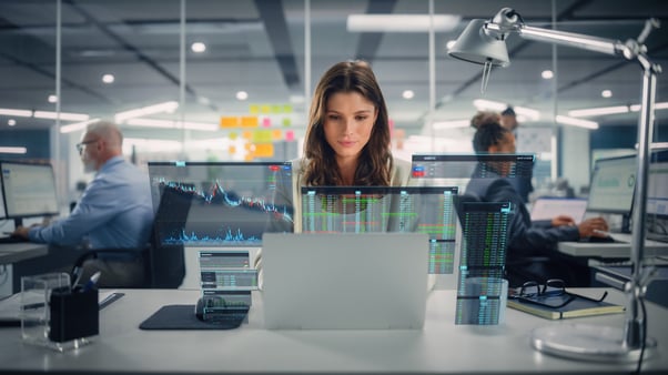 Profissional trabalhando em notebook em escritório com monitores exibindo gráficos